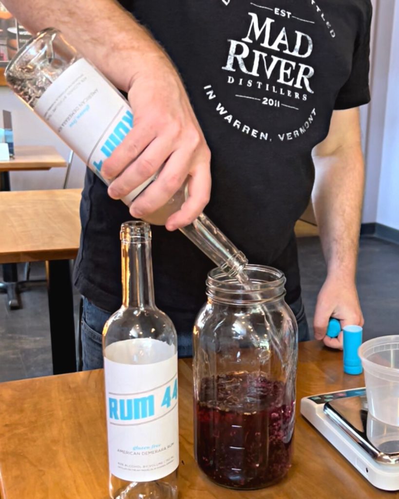 A man is pouring a bottle of Rum 44 into a tall glass mason jar filled with hibiscus flowers.