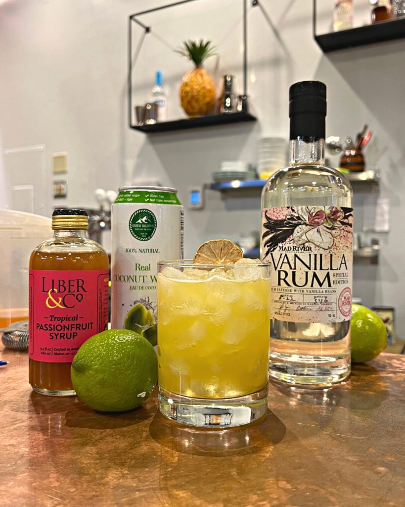 Fat-Washed Vanilla Tropics cocktail next to a bottle of Vanilla Rum, Coconut Water, passionfruit syrup and a lime